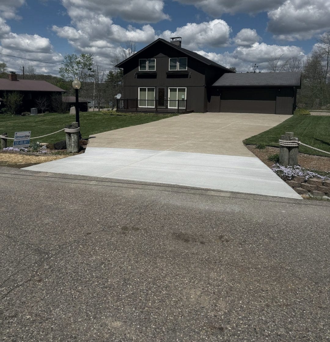 The finished concrete driveway and apron in front of a brown two-story home, broom-swept and cured, meeting the road under a clouded sky.