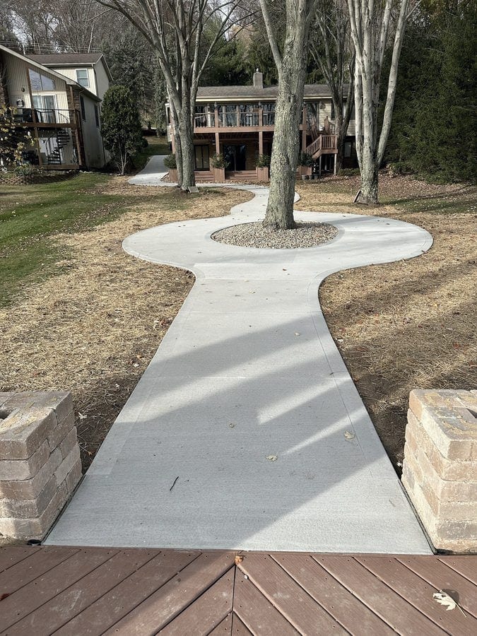 A curved concrete walkway runs between mature trees from a deck to a home, with tree rings cut into the walk and brick pillars at the near end.