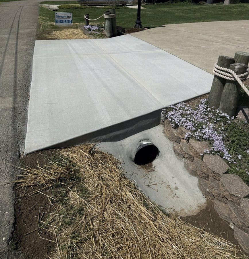 The finished concrete drive apron poured over the culvert, with the pipe end and the dry-laid stone headwall left in place and creeping phlox in bloom alongside.
