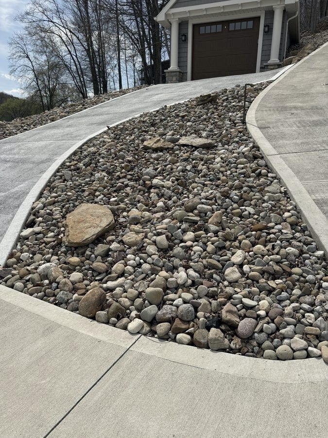 A sweeping river rock bed with a flat sandstone accent stone curves between two runs of new concrete drive in front of a garage.