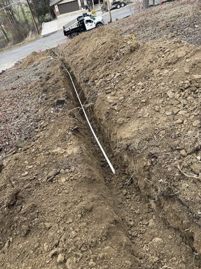 A long, narrow drain trench with white pipe laid in runs down a bare hillside toward a road where a Streamline dump truck is parked.