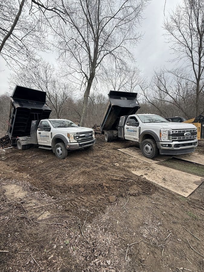 Two Streamline dump trucks with their beds raised tip loads onto a freshly cleared lot, with plywood mats laid down over the soft ground.