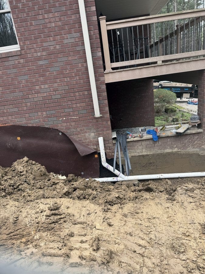 Foundation drainage board and white pipe are set against a brick wall under a raised deck, with a Streamline dump trailer parked beyond.