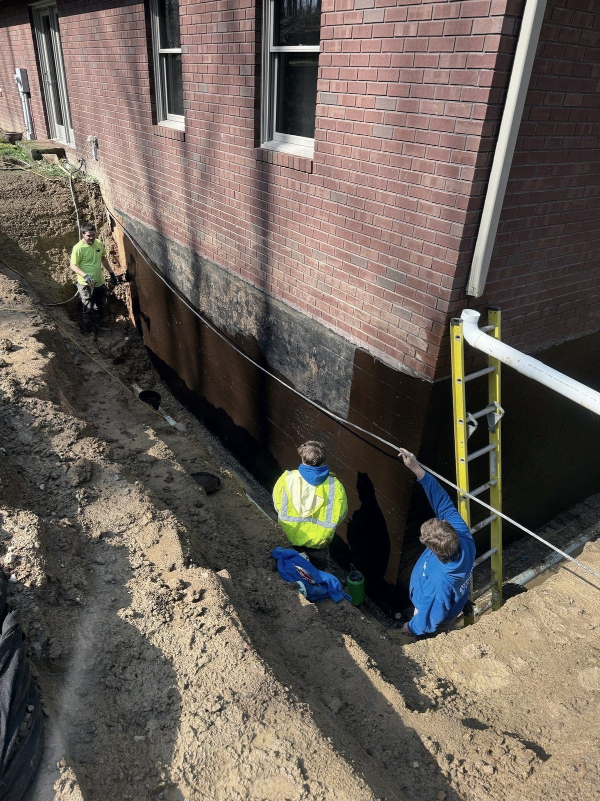 Two of the crew work in an open trench along a brick foundation that has been coated with brown waterproofing membrane, with a ladder and a level line in place.
