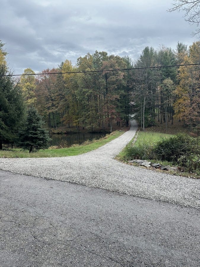A finished gravel drive curves away from an asphalt road and into fall woods, with a pond visible through the trees.