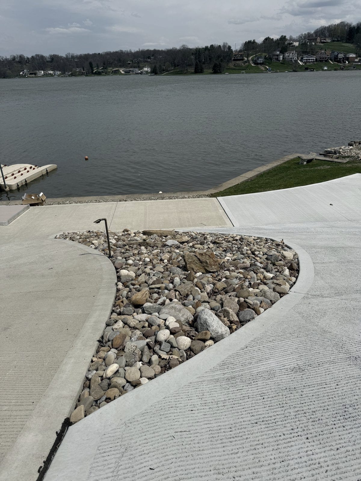 A teardrop island of rounded river rock set into a broom-finished concrete drive that runs down to a lake, with a floating dock and the far shore beyond.
