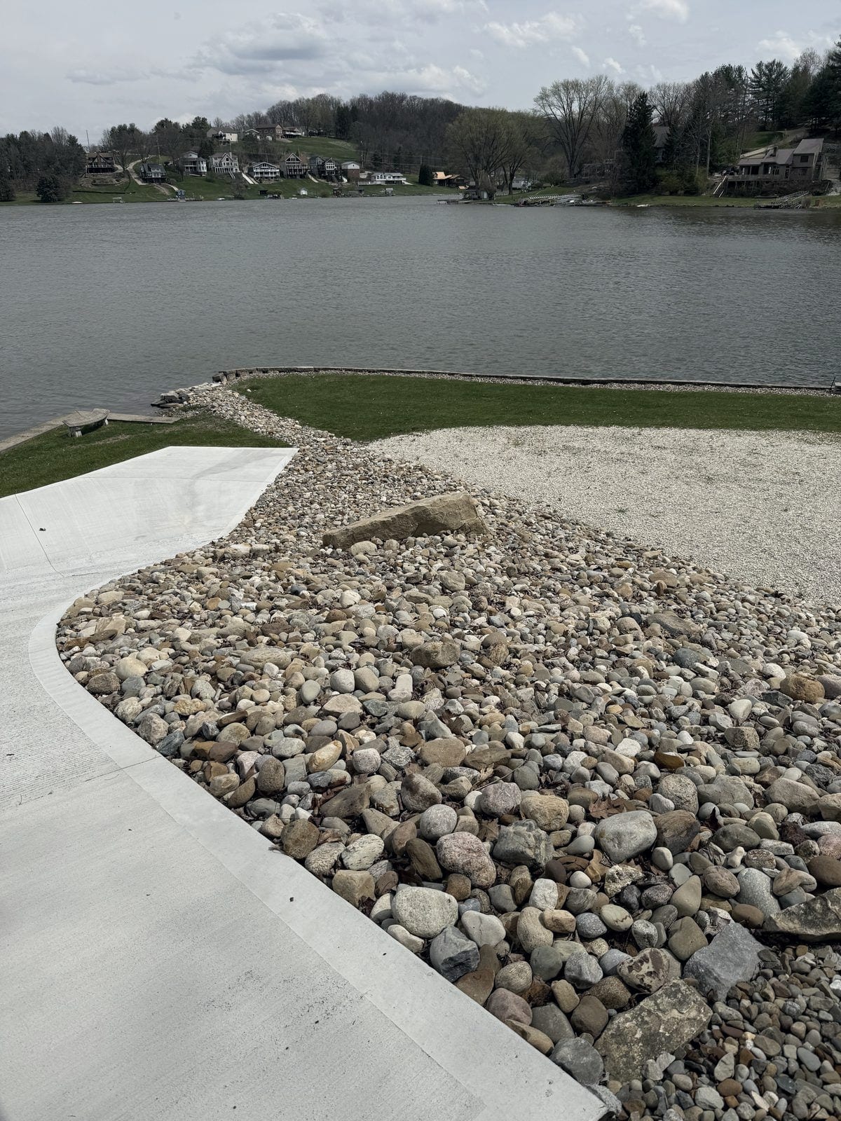 A finished concrete drive and a wide bed of rounded river rock meet a new lawn that runs down to a stone-edged lake shoreline, with wooded hills and lake houses across the water.