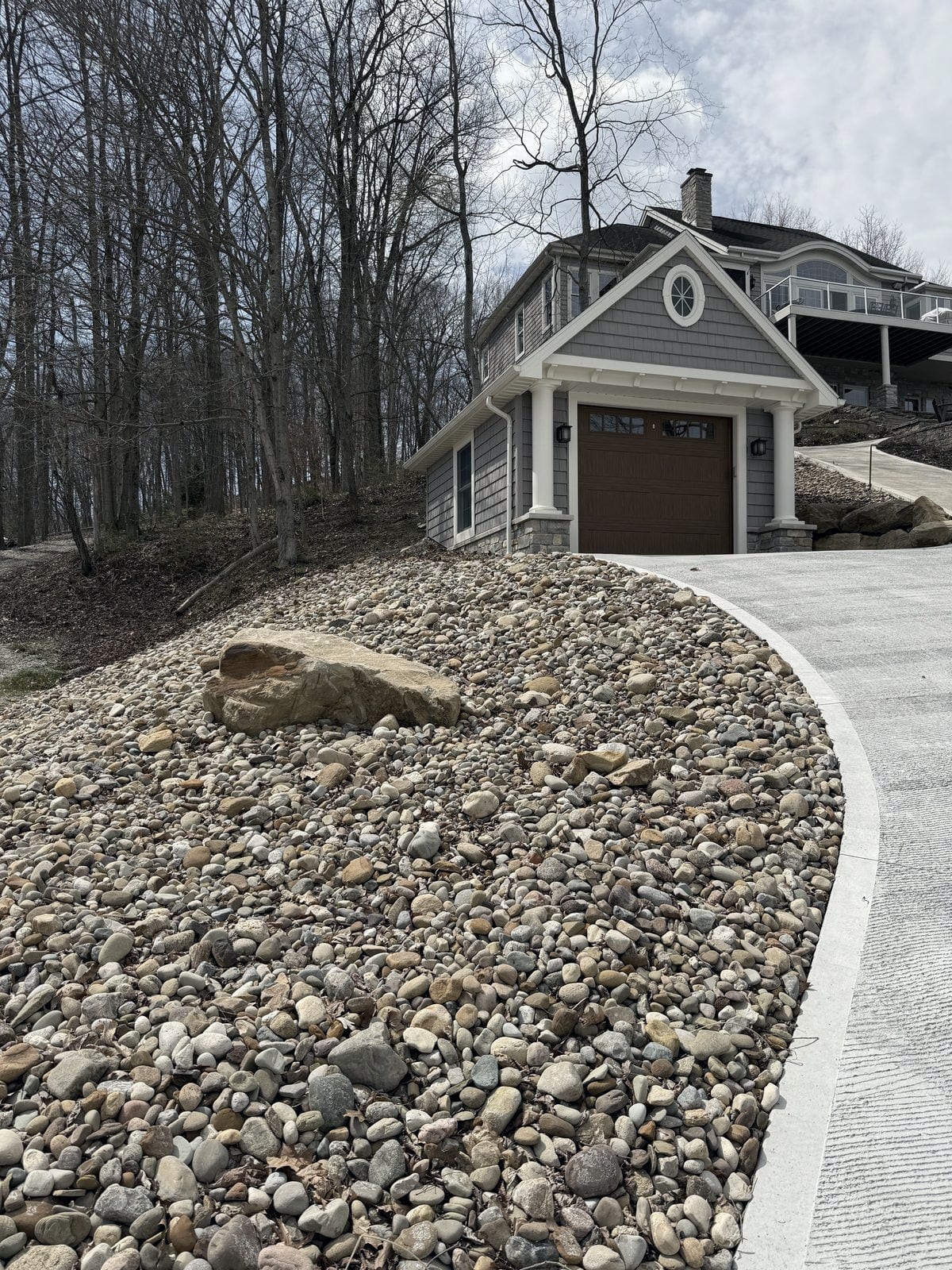 A river rock bank with a set sandstone boulder runs the length of a new concrete drive beside a shingled lakeside garage on a wooded slope.