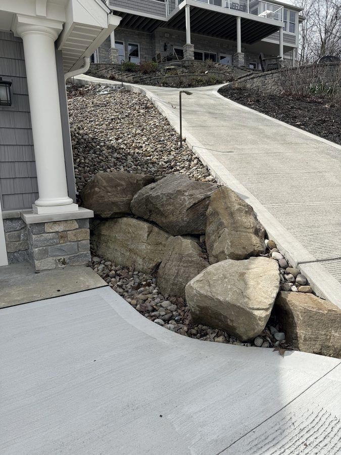 Stacked sandstone boulders and river rock hold a planted slope between a stone-based porch column and a new concrete walk.