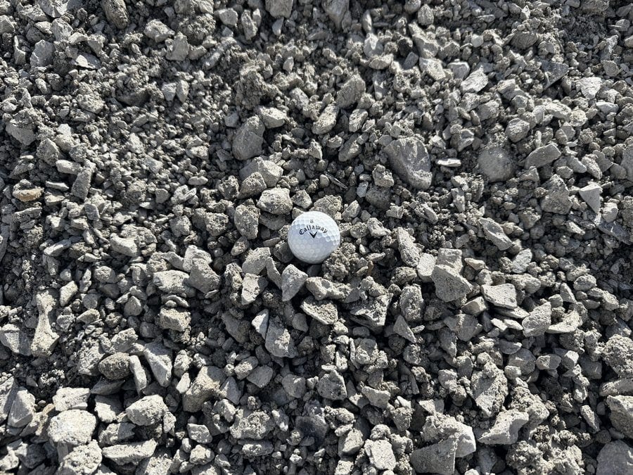 Gray crushed limestone with dark fines mixed through it, shown with a golf ball for scale.