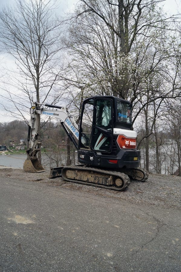 A Streamline-decaled mini excavator parked on a drive above a lake, with the water visible through the bare trees.