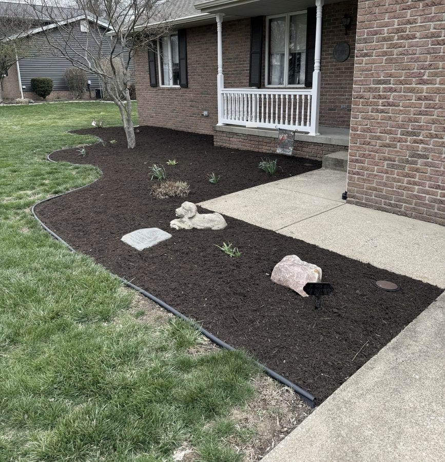 A wide brown mulch bed follows the concrete front walk of a brick ranch home, curving out around a tree and a set of accent stones.