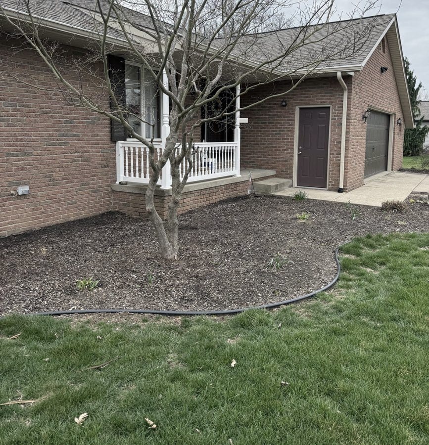 A large brown mulch bed sweeps around a bare tree across the front of a brick ranch home with a white porch rail.