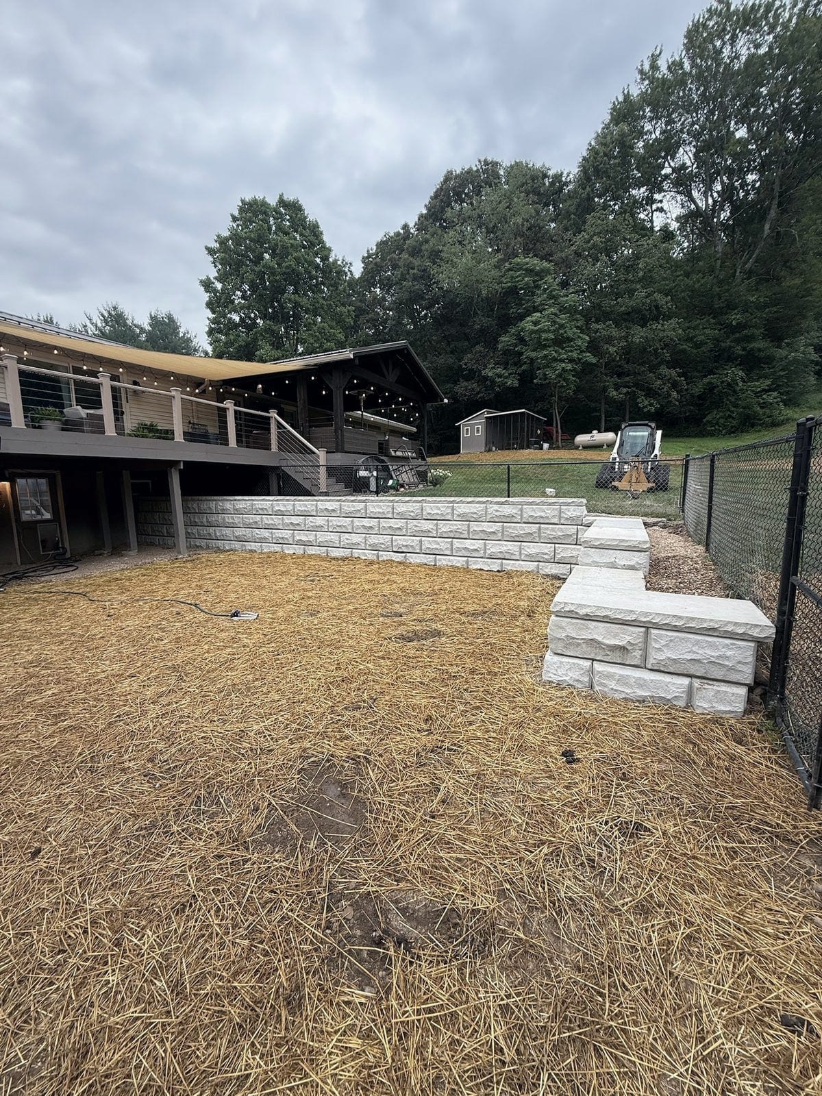 A two-tier gray block retaining wall with a wide stone step terraces a back yard that has been graded and covered with straw, beside a fenced lawn and a covered pavilion.