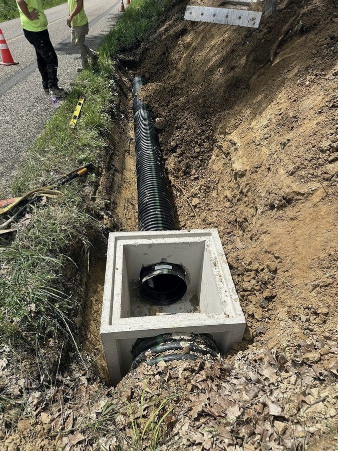 Black corrugated culvert pipe is bedded in a trench cut into a roadside bank, with a level and traffic cones set out on the pavement alongside.