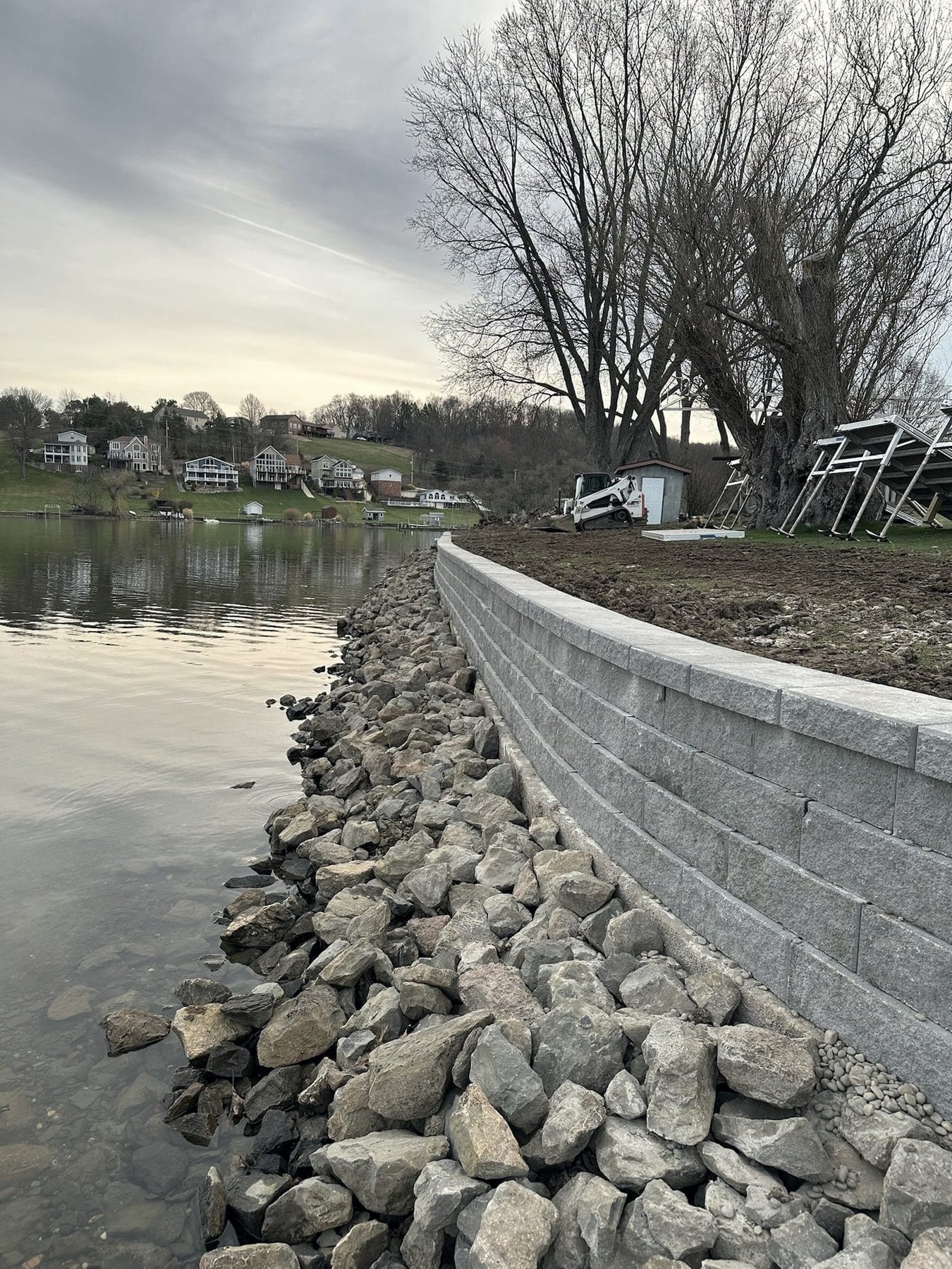 A finished gray block sea wall curves along a lake shoreline, with a bank of large riprap stone stacked at the waterline in front of it and lake houses on the far shore at dusk.