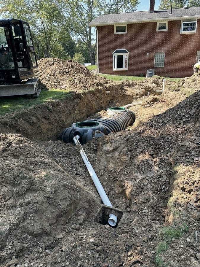 A septic tank set in an open trench with its inlet pipe run out along the bottom of the excavation, beside a brick home.