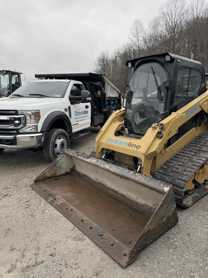 A Streamline-decaled tracked skid steer with its bucket lowered, parked beside one of the dump trucks.