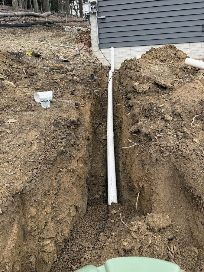 A white drain line and a catch basin box are set in an open trench alongside a home with gray siding.