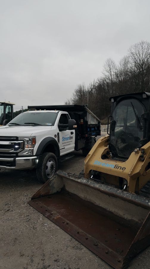 A Streamline dump truck and a tracked skid steer parked side by side on a gravel lot at the edge of the woods.